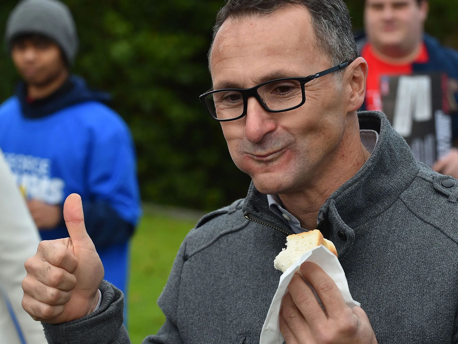 Man in glasses giving a thumbs up while eating a sandwich, with a group of people in the background.