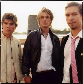 Three men stand outdoors; the center one wears a black jacket, the others in light and dark suits. Background shows a tower.