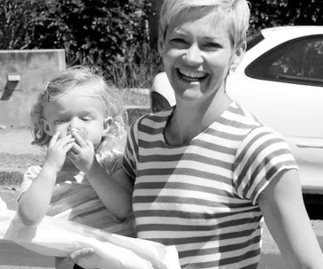 Smiling woman in a striped shirt holding a child who is rubbing their nose, both in black and white.
