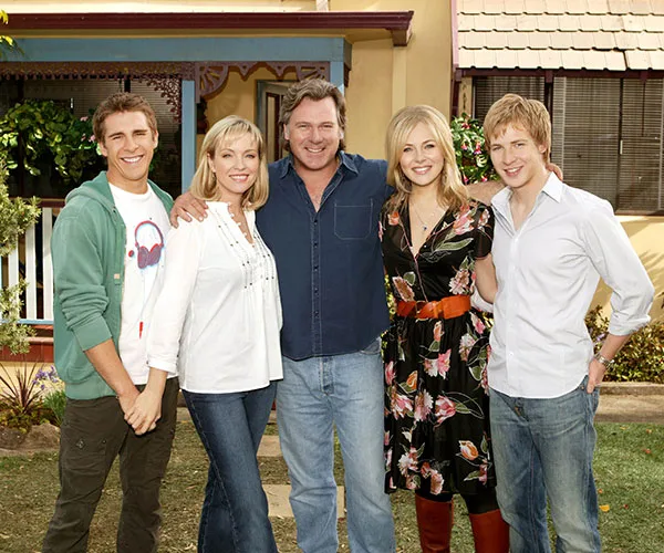 A group of five people smiling, standing in front of a house. They appear casually dressed and happy.