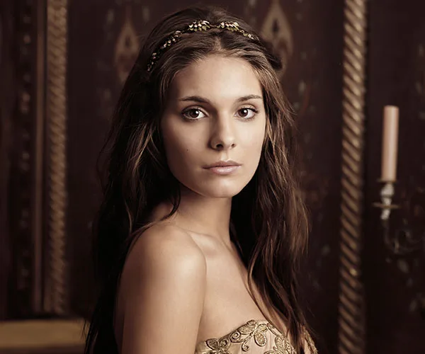 Young woman with long brown hair and a jeweled headband dressed in medieval-inspired attire, standing indoors.