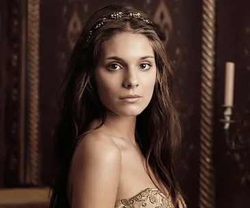 Young woman with long brown hair and a jeweled headband dressed in medieval-inspired attire, standing indoors.