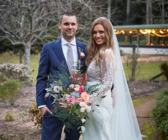 Bride in lace wedding dress and groom in navy suit smiling outdoors, holding a colorful bouquet.