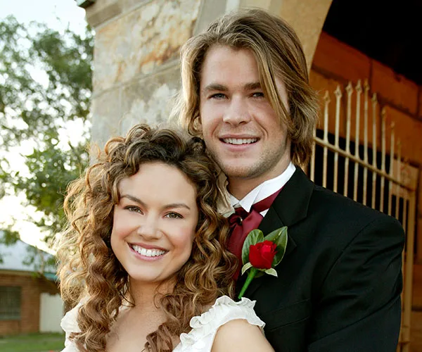 A smiling couple at a formal event, with the man in a suit and red rose, and the woman in a white dress.