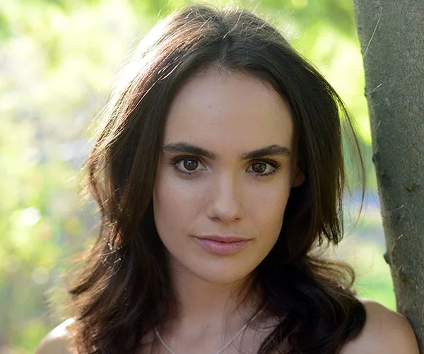 A woman with long dark hair stands outdoors, sunlight highlighting her face against a blurred natural background.