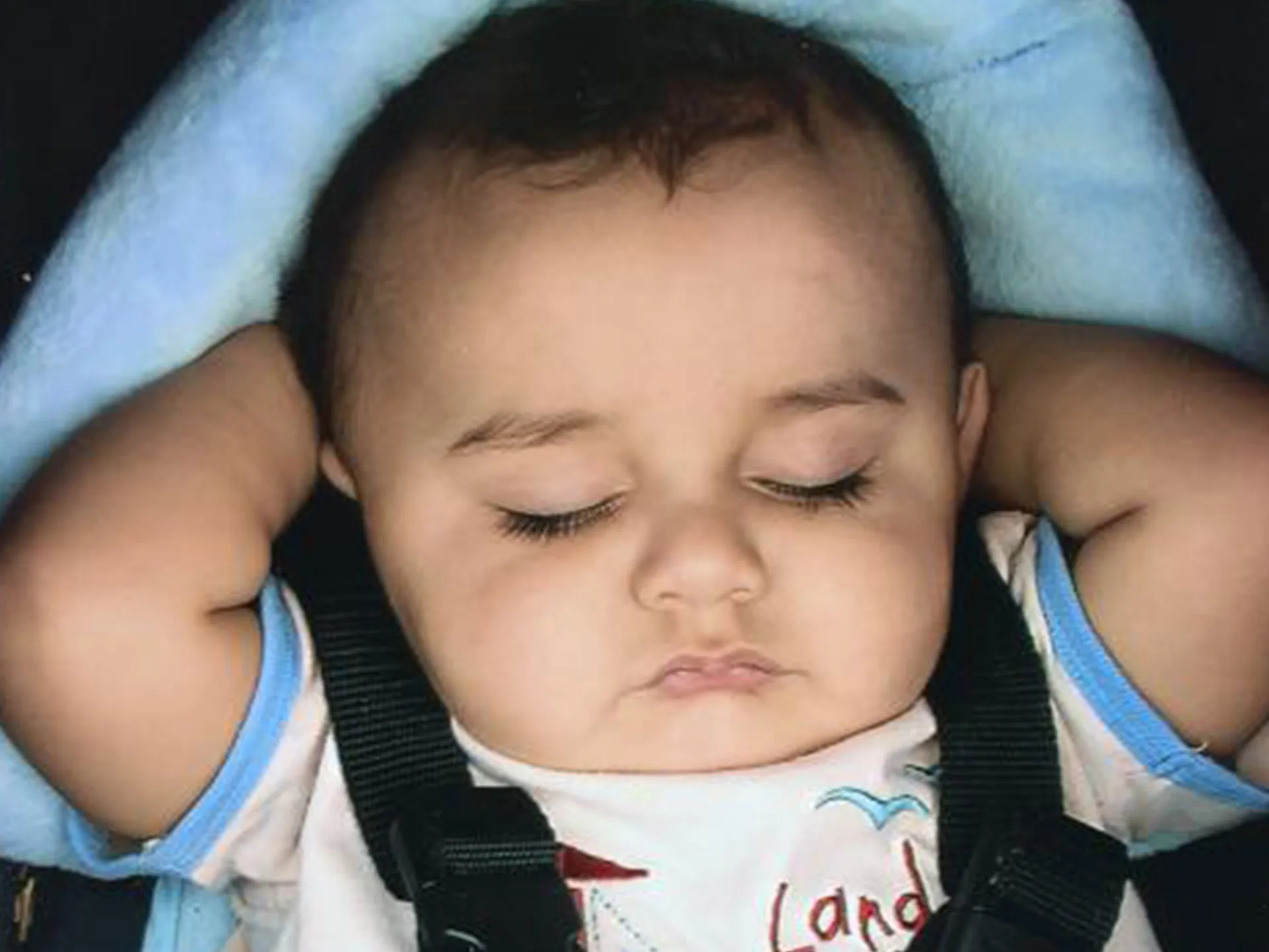 A sleeping baby with raised arms, wearing a white shirt in a car seat with a blue blanket.