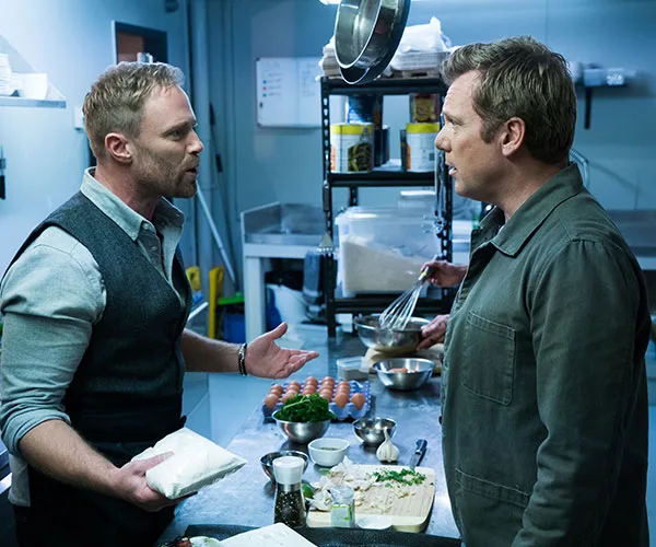 Two men having a discussion in a kitchen, with cooking ingredients visible on the counter.