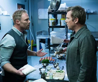 Two men having a discussion in a kitchen, with cooking ingredients visible on the counter.