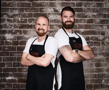 Two men in MKR aprons stand back-to-back, smiling with arms crossed in front of a brick wall.
