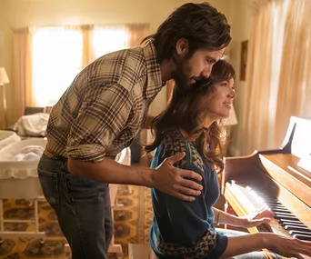 A man lovingly embraces a woman playing a piano in a warmly-lit room with a crib in the background.