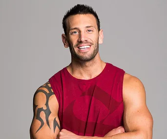 Man in a sleeveless red shirt with arm tattoo smiling confidently, arms crossed against gray background.