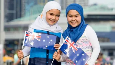 Two young girls wearing hijabs, smiling and holding Australian flags outdoors.