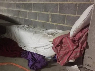 Sleeping area with makeshift bedding on a concrete floor in a corner, with pillows and blankets.