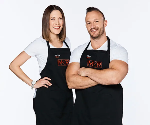 Two "My Kitchen Rules" contestants wearing black aprons with the show's logo, smiling and posing against a white background.