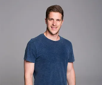 Young man in a blue t-shirt smiling, standing against a neutral gray background.