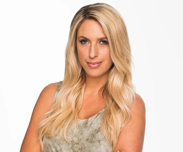 A person with long blonde hair wearing a sleeveless top, looking confidently into the camera, set against a plain background.