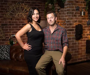 A couple sits together on a leather couch in a cozy, warmly lit living room with exposed brick walls and decorative art.