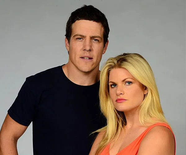 Two actors from "Home and Away" pose together against a plain background, one in a navy shirt, the other in an orange top.