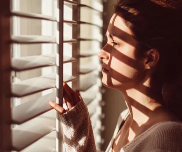 A woman in soft light looks through window blinds, casting striped shadows across her face.