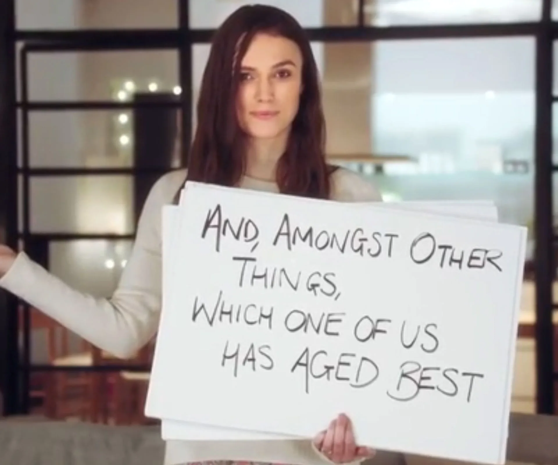 Woman holding a sign saying, "And, amongst other things, which one of us has aged best," in a room with lights.