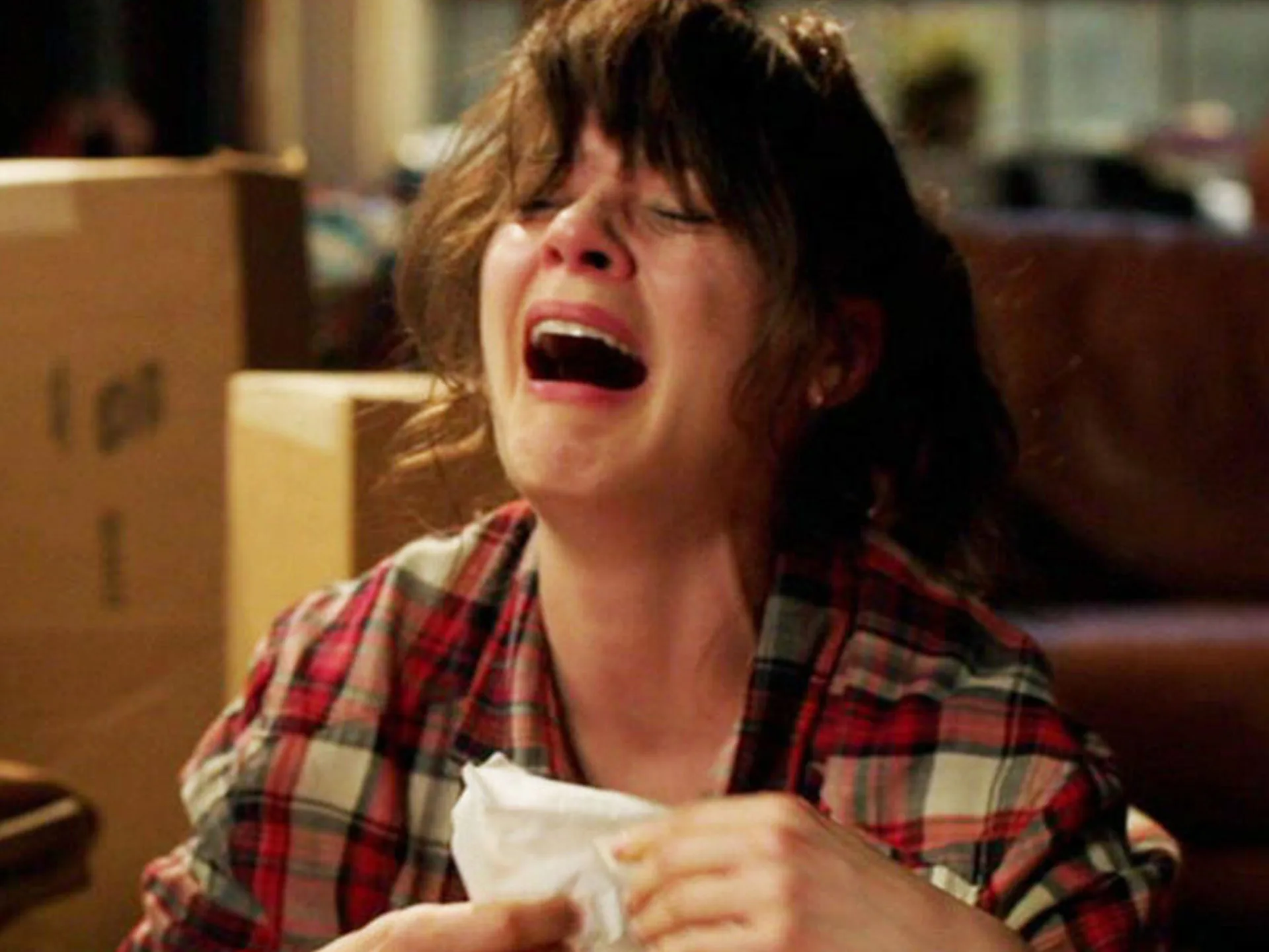 Woman in a plaid shirt crying intensely while holding tissues, surrounded by boxes in a room.