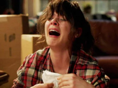 Woman in a plaid shirt crying intensely while holding tissues, surrounded by boxes in a room.
