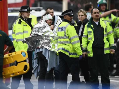 Emergency responders assist an injured person wrapped in a foil blanket, surrounded by police and medical personnel.