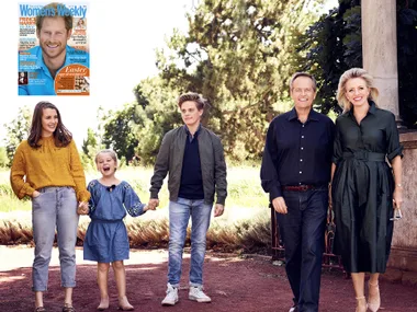 A family standing together outdoors, smiling in casual attire, with trees in the background; magazine cover inset featuring Prince Harry.
