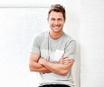 Man in a grey t-shirt smiling, arms crossed, relaxed pose against a light backdrop.