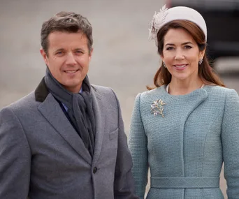 Two people dressed in formal attire, one in a grey suit and scarf, the other in a teal outfit with a hat and brooch.