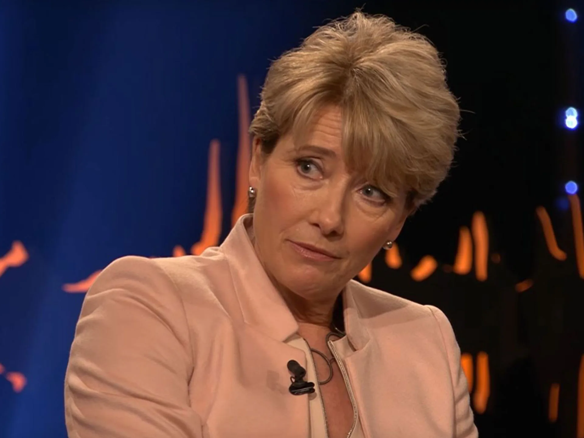 A person in a pink blazer speaks on a stage, with an orange and blue background.