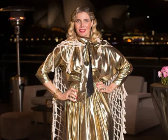 A woman in a shiny gold outfit poses confidently outdoors at night, with a city skyline background.
