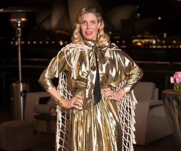 A woman in a shiny gold outfit poses confidently outdoors at night, with a city skyline background.
