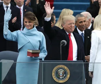 Melania Trump in blue coat, standing next to Donald Trump at an inauguration event, both raising hands.