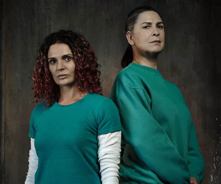Two characters from Wentworth Season 4 in teal uniforms stand together against a dark background.