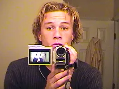 Person with a camcorder filming a mirror reflection, wearing a dark sweater.