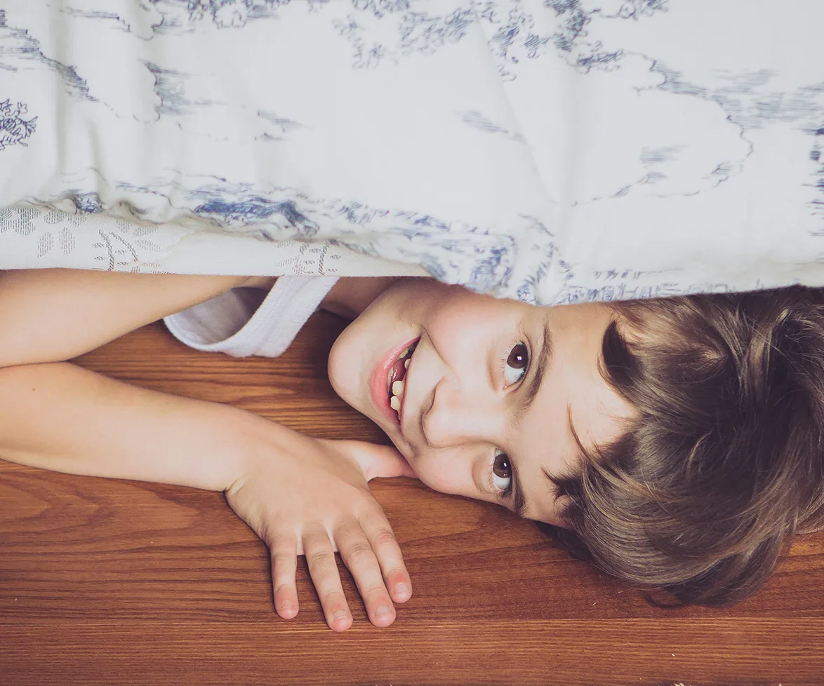 A smiling boy lies under a bed with wooden flooring, partially covered by a patterned blanket.