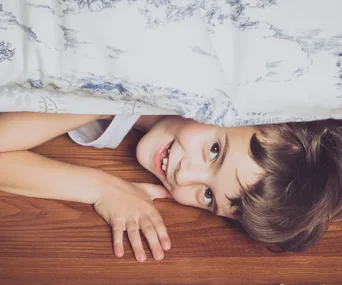 A smiling boy lies under a bed with wooden flooring, partially covered by a patterned blanket.