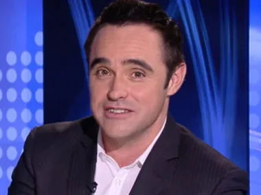 Television presenter in a suit, seated in a studio with a blue background displaying digital patterns.