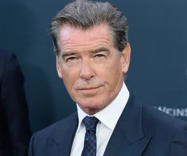 Pierce Brosnan in a suit, looking directly at the camera, with a solid-colored background.