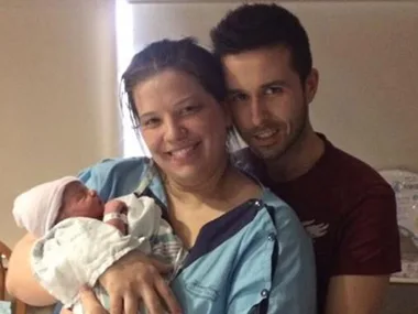 A woman in hospital gown holding a newborn baby, smiling with a man standing behind her.