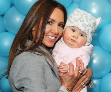 Smiling woman holds a baby wearing a fluffy, patterned hat, against a backdrop of blue balloons.