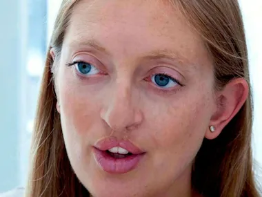 A woman with long hair and blue eyes, wearing a small earring, looking thoughtful.