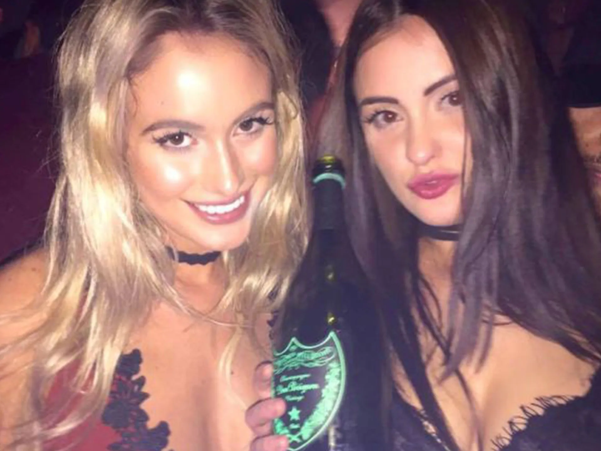 Two women smiling and posing with a bottle at a nighttime event, both wearing choker necklaces.