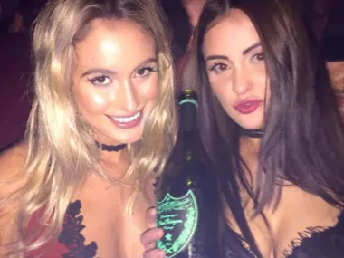 Two women smiling and posing with a bottle at a nighttime event, both wearing choker necklaces.