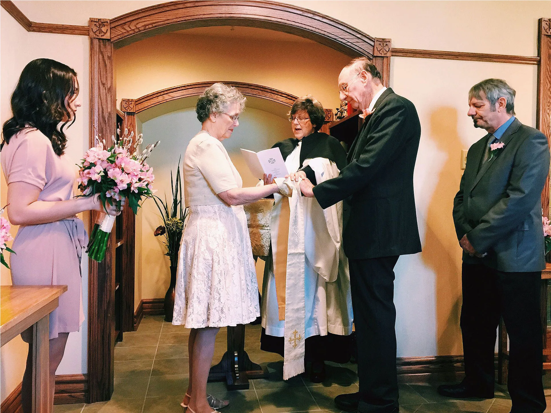 Elderly couple exchanging vows at a wedding ceremony with a bouquet-holding bridesmaid and witnesses standing by.