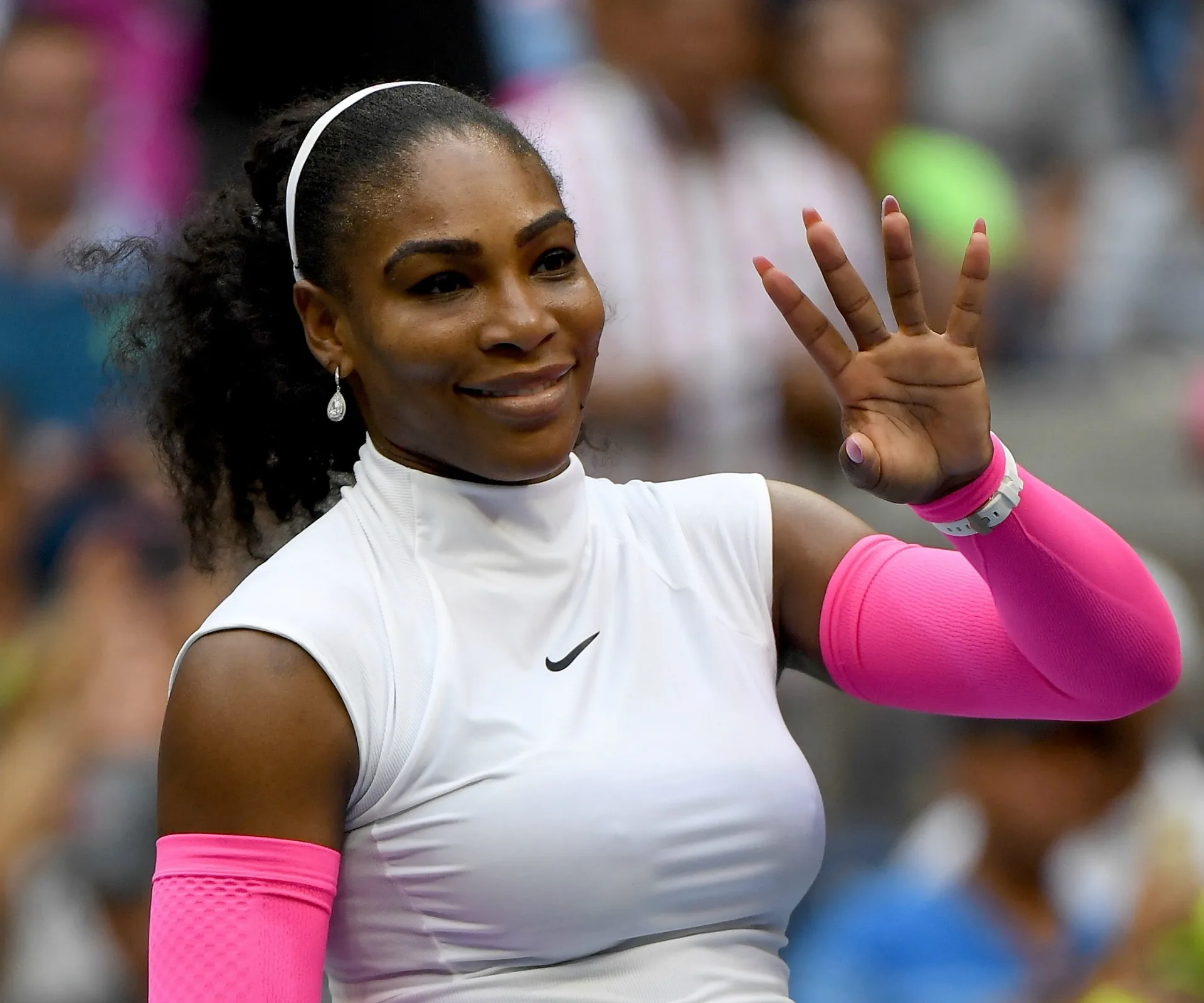Tennis player in a white top and pink sleeves waves to the crowd, smiling.