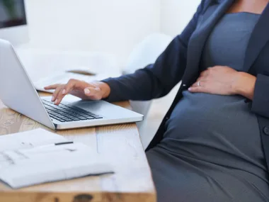 Pregnant woman in business attire working on a laptop at a desk.