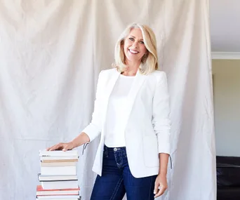 A woman in a white blazer and blue jeans stands smiling next to a stack of books against a light-colored backdrop.