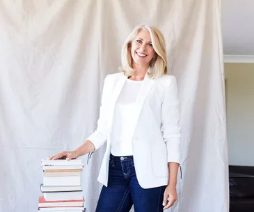 A woman in a white blazer and blue jeans stands smiling next to a stack of books against a light-colored backdrop.